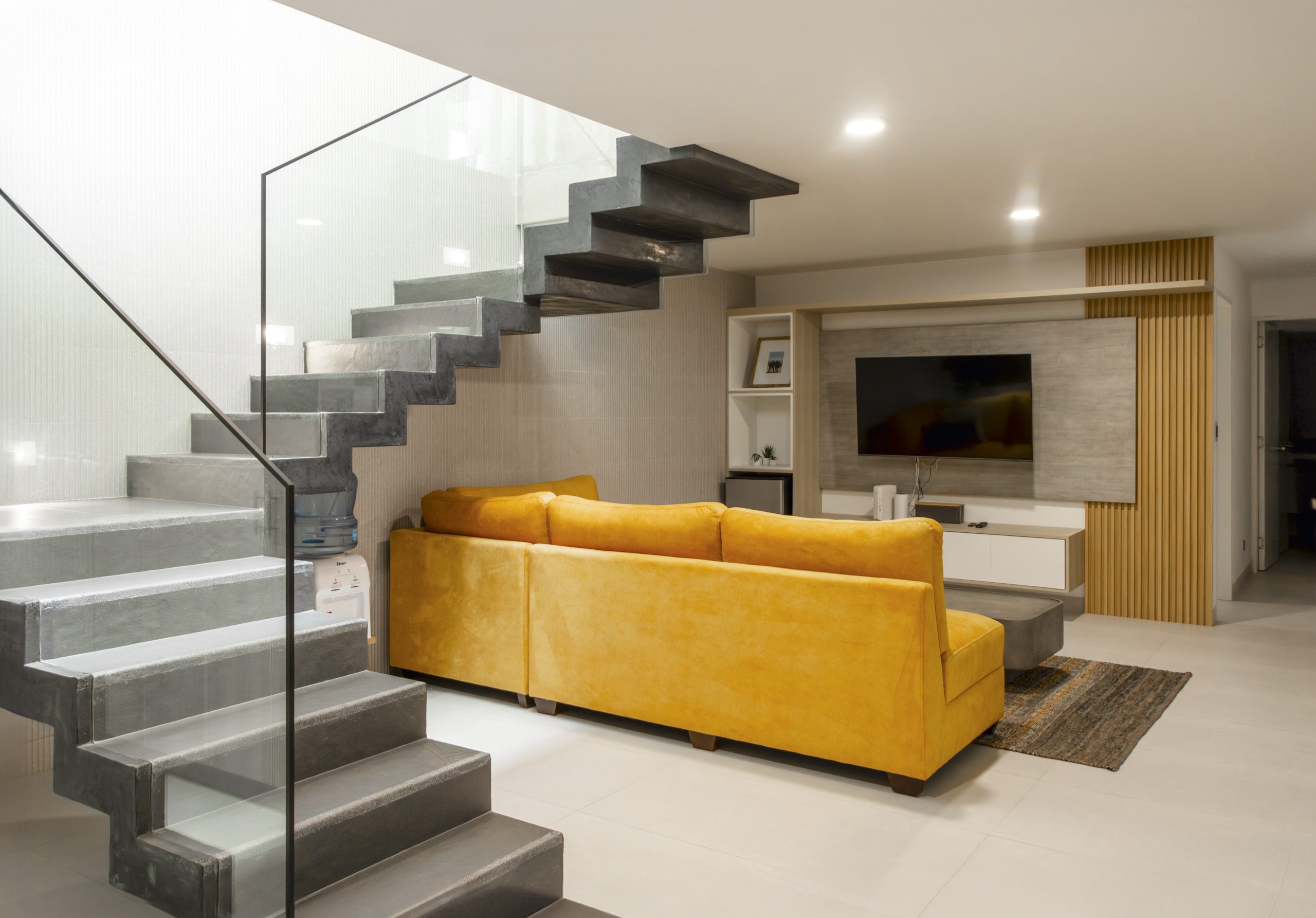La escalera de concreto y vidrio convive con un living funcional, donde el sofá mostaza introduce calidez en un ambiente de líneas limpias y tonos neutros.