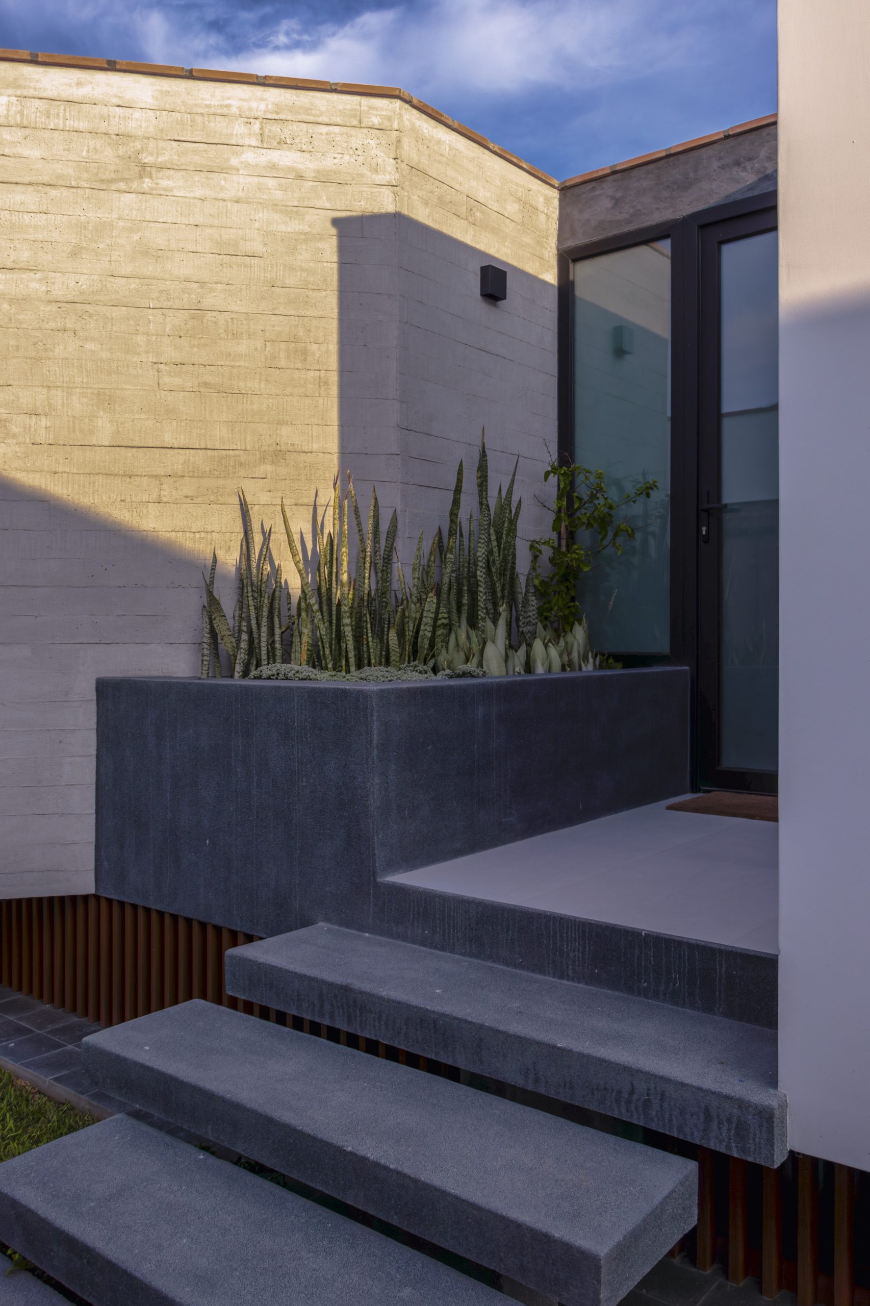 El acceso a la vivienda se resuelve con recorridos claros, luz natural y vegetación integrada, marcando desde el inicio una experiencia calmada y contenida.