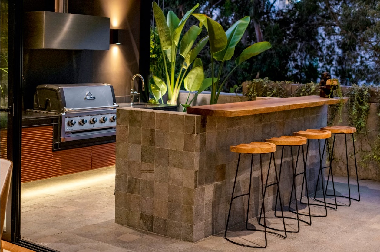 La barra y el área de parrilla, revestidas en piedra talamoye, articulan la terraza como un espacio pensado para cocinar, reunirse y prolongar la vida social al aire libre.